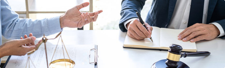 Two lawyers at a desk discuss negotiate while taking notes.