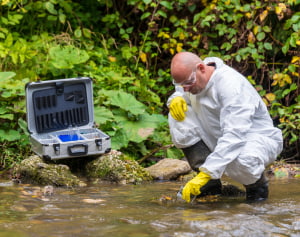 A researcher in protective clothing kneels beside a fresh water source collecting a sample to test for water contamination.